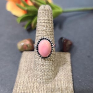 May include: A silver ring featuring a pink oval gemstone set in a beaded bezel. The ring is displayed on a fabric ring holder.
