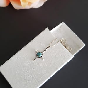 May include: A silver necklace with a turquoise cross pendant in a white box with a textured pattern.