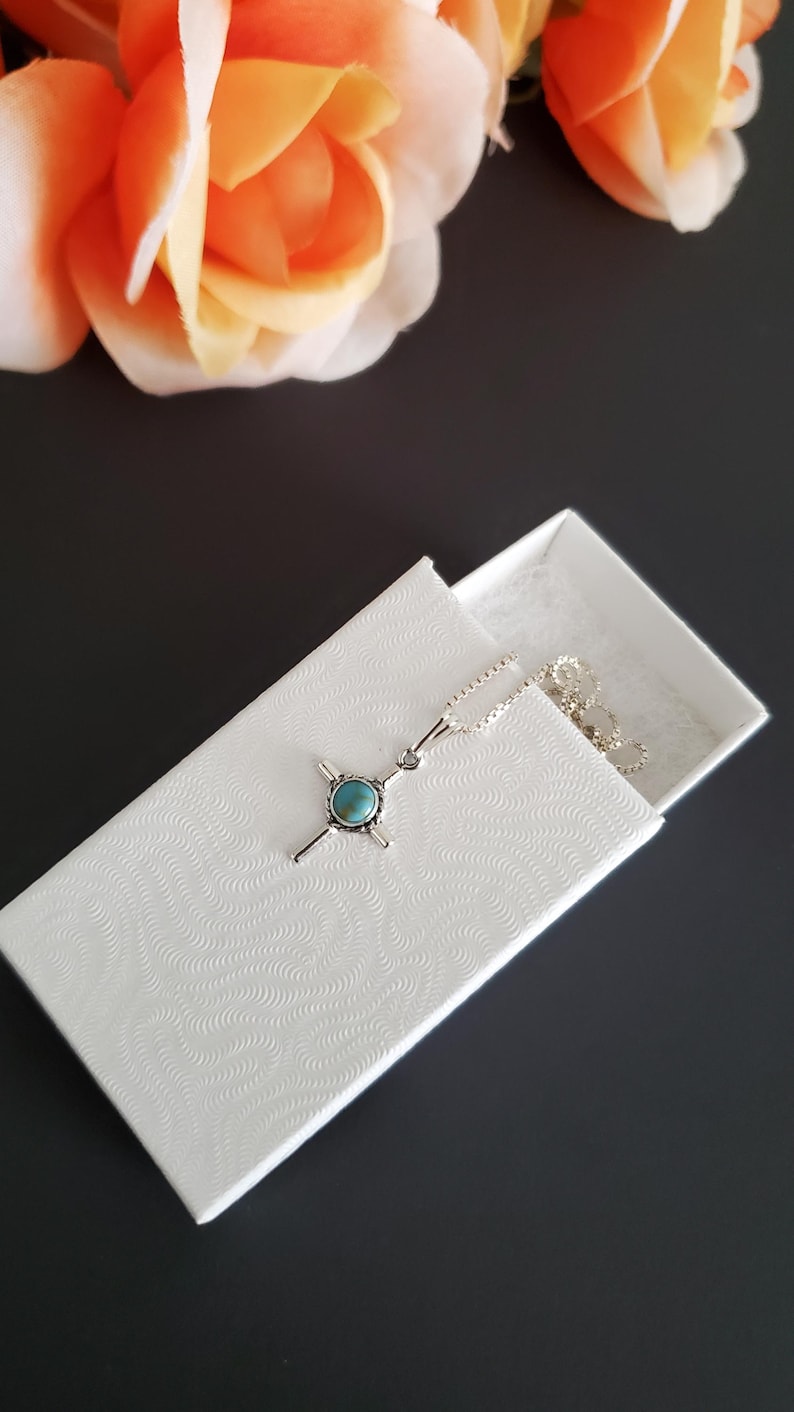 May include: A silver necklace with a turquoise cross pendant in a white box with a textured pattern.