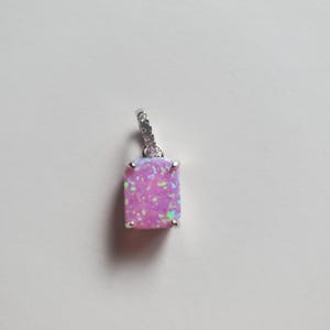 Puede incluir: Un colgante de plata con una piedra preciosa de ópalo rosa. El ópalo tiene forma rectangular y está engastado en un engaste de plata. El colgante tiene una anilla de plata para sujetarlo a una cadena.