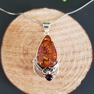 Puede incluir: Un collar con colgante de plata con una piedra de ámbar en forma de lágrima. El ámbar tiene un tono marrón dorado cálido con patrones internos. Una pequeña gema roja oscura está engastada debajo del ámbar.