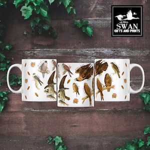 (All 3 sides of mug) A white ceramic mug featuring labelled vintage art of carnivorous birds of prey. These include a hobby, peregrine falcon, osprey, 
kestrel, merlin, golden eagle, red kite, buzzard and sparrowhawk. Water colour feathers and flowers are arranged around the birds.