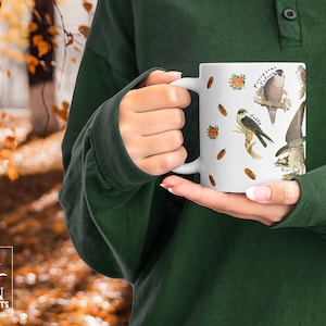 A person in a forest holding a white ceramic mug. The cup features labelled vintage art of carnivorous birds of prey. These include a hobby, peregrine falcon, osprey, kestrel, merlin, golden eagle, red kite, buzzard and sparrowhawk. Water colour feathers and flowers are arranged around the birds.