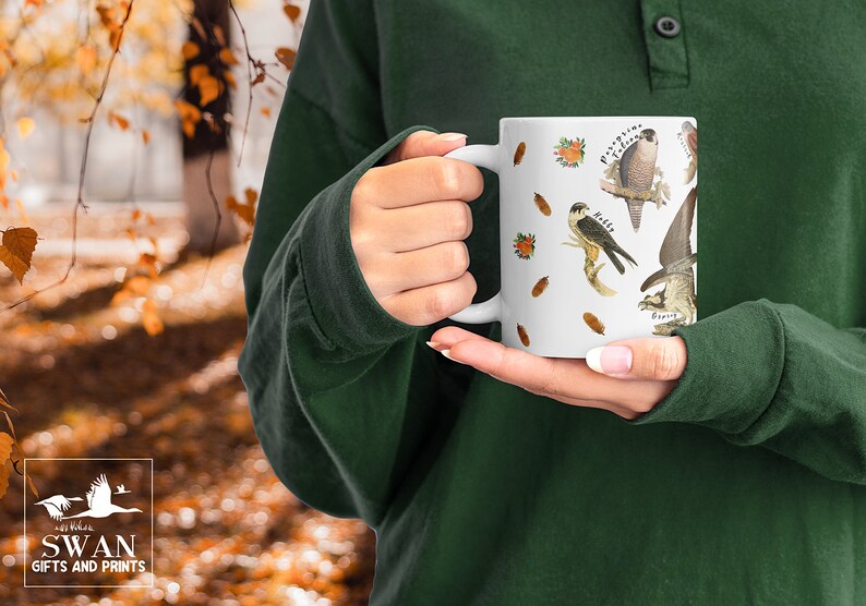 A person in a forest holding a white ceramic mug. The cup features labelled vintage art of carnivorous birds of prey. These include a hobby, peregrine falcon, osprey, kestrel, merlin, golden eagle, red kite, buzzard and sparrowhawk. Water colour feathers and flowers are arranged around the birds.