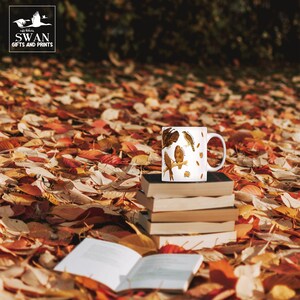 A white ceramic mug on a pile of books in the forest. The cup features labelled vintage art of carnivorous birds of prey. These include a hobby, peregrine falcon, osprey, kestrel, merlin, golden eagle, red kite, buzzard and sparrowhawk. Water colour feathers and flowers are arranged around the birds.
