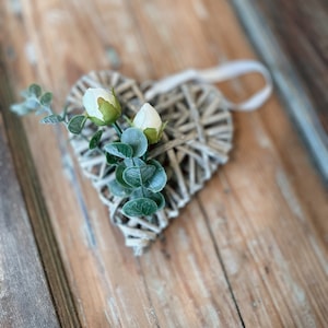 May include: A woven heart-shaped decoration with a cream-colored ribbon for hanging. The heart is adorned with white flower buds and green leaves. The background is a rustic wooden surface.