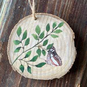 May include: A wooden round ornament with a painted image of a butterfly and green leaves. The ornament has a twine string for hanging.