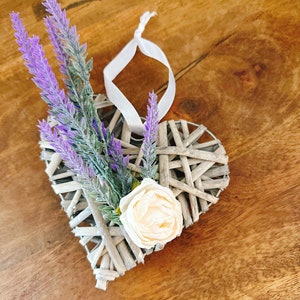 May include: A woven heart-shaped decoration with lavender and a cream-colored rose. The heart is made of light gray woven material and has a white ribbon for hanging. The lavender is purple and green.