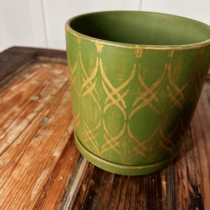 May include: A green ceramic planter with a matching saucer. The planter features a gold geometric pattern. The planter is sitting on a wooden surface. The planter is approximately 6 inches in diameter and 5 inches tall.