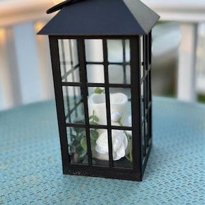 Puede incluir: Linterna de metal negro con un diseño de paneles de vidrio. La linterna tiene un arreglo floral blanco en su interior. La linterna está sentada sobre una mesa tejida azul.