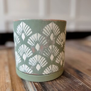 May include: A cylindrical, sage green ceramic planter with a matching saucer. The planter is decorated with a repeating white fan-shaped pattern. The rim of the planter has a natural terracotta color.