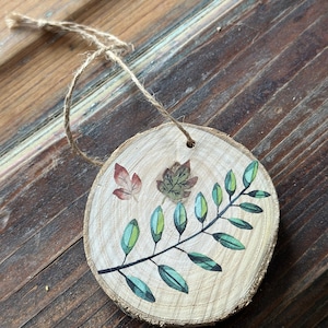 May include: A wooden ornament with a twig and leaves painted on it. The ornament is brown and has a natural wood grain. There are also three small leaves painted in red, orange, and yellow.