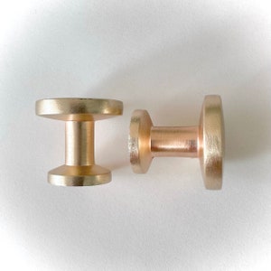 May include: Two gold-toned metal cabinet knobs. One knob is round with a cylindrical base. The other knob is round with a short, cylindrical base.