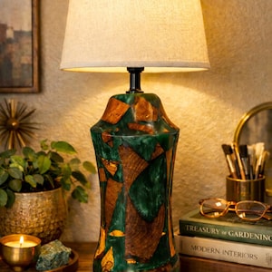 May include: A table lamp with a beige lampshade. The base is made from green resin and wood. The lamp is on a wooden surface, with books and other decorative items visible. The lamp's design is unique.