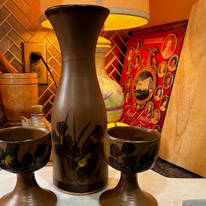 Może przedstawiać: Zestaw trzech brązowych ceramicznych naczyń do picia z kwiatowym wzorem. Zestaw zawiera wysoki dzbanek i dwie mniejsze filiżanki ze stopkami.