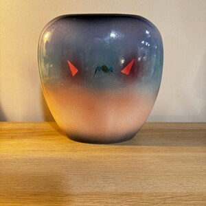 May include: A ceramic vase with a colour gradient, shifting from dark blue at the top to a soft pink at the base. The vase has abstract red and teal accents, providing a unique artistic element. The vase is displayed on a wooden surface.