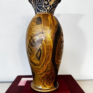 Könnte beinhalten: Eine dekorative Keramikvase mit breitem Boden und ausgestelltem Oberteil. Die Vase hat ein braunes und goldenes Wirbelmuster auf dem Körper und einen schwarzen Oberteil mit weißen geometrischen Designs. Sie steht auf einem roten Buch.