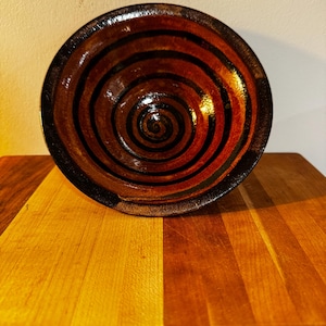May include: A handmade ceramic bowl with a spiral design. The bowl features concentric circles in shades of brown, red, and black. The bowl is sitting on a wooden surface.