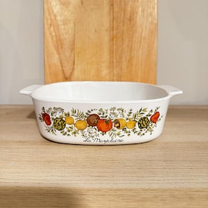 Pode incluir: Travessa oval branca com um design colorido de vegetais. A travessa apresenta uma borda de tomates, limões e outros produtos. As palavras "La Marjolaine" estão impressas na frente. Uma tábua de corte de madeira está no fundo.