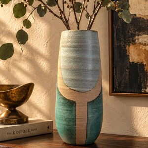 May include: A tall, cylindrical ceramic vase featuring a textured surface with a color-blocked design. The upper portion is a light blue, while the lower section is a deep teal, both separated by a vertical and curved band of unglazed beige clay.