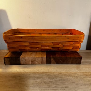 May include: An orange rectangular woven basket sits on a wooden chopping board with light and dark brown sections. The basket has a rounded rim and is made of woven wood strips.