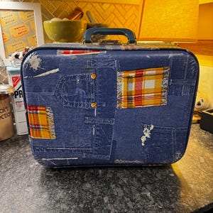 Pode incluir: Uma mala azul vintage com um design de patchwork de ganga. A mala apresenta bolsos de ganga falsos e detalhes em tecido xadrez em amarelo, vermelho e branco. Possui uma pega azul escura e ferragens douradas. A mala está sobre uma bancada escura.