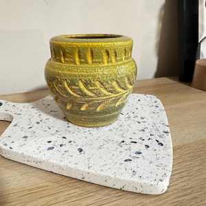 Pode incluir: Um pequeno vaso redondo de cerâmica com um padrão texturizado em forma de folha em tons de verde e amarelo. O vaso está sobre uma tábua de cortar branca salpicada com uma borda arredondada.
