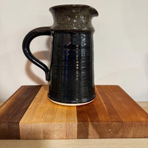 May include: A handmade, dark blue ceramic pitcher with a textured surface and a dark brown speckled rim. The pitcher has a curved handle and a spout for pouring. It sits on a wooden cutting board with various wood tones.