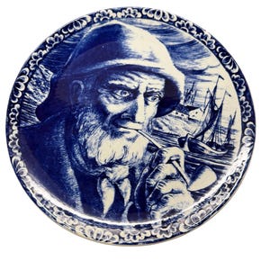 Pode incluir: Prato decorativo azul e branco com uma ilustração detalhada de um homem idoso com barba, chapéu e cachimbo. O fundo mostra veleiros e o mar. O prato tem uma borda decorativa.