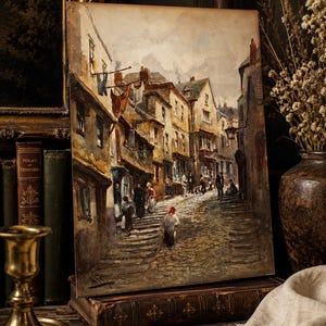 May include: An oil painting on a rectangular canvas depicting a narrow, cobblestone street lined with multi-story buildings in muted earth tones. Several figures walk along the path, and laundry hangs from lines between the structures.