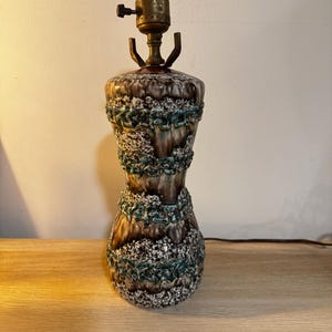 May include: A vintage ceramic table lamp with a textured, hourglass-shaped body. The lamp features a brown, white, and teal color scheme. The lamp's brass-colored socket and switch are visible at the top. The lamp is approximately 14 inches tall.