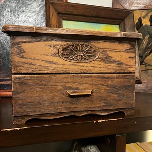 Peut inclure: Une petite boîte en bois avec un seul tiroir et un motif de tournesol sculpté sur le devant. La boîte est brune et a une finition vieillie.