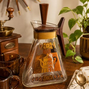 Könnte beinhalten: Eine klare Glaskaraffe mit dreieckigem Boden, braunem Kunststoffdeckel und Griff, einem goldfarbenen Metallhalsband sowie einem dekorativen orange-goldenen Grafikdruck einer Teekanne und Blumenmotiven auf der Vorderseite.