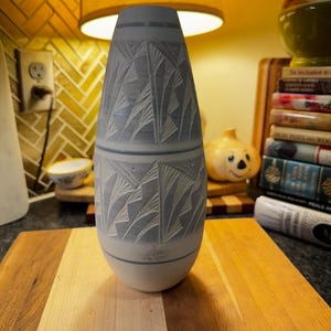 Może przedstawiać: Jasnoniebieski ceramiczny wazon z rzeźbionym wzorem stylizowanych liści. Wazon jest cylindryczny z lekko rozszerzonym górnym i dolnym brzegiem. Stoi na drewnianej desce do krojenia. Wysokość ok. 25 cm.