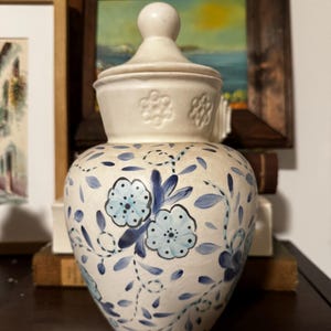 Puede incluir: Un jarrón de cerámica blanco con tapa, adornado con motivos florales azules. El jarrón tiene forma bulbosa y una tapa a juego con un pomo. El diseño floral incluye flores y hojas azules. El jarrón se utiliza probablemente para almacenamiento decorativo.