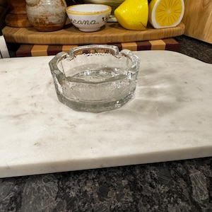 Puede incluir: Un cenicero de vidrio transparente con una superficie texturizada. El cenicero es redondo y tiene un borde ligeramente elevado.