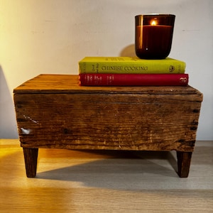 May include: A small, rustic wooden table with four legs. On top, there are two books, one yellow and one red, and a lit brown glass candle. The yellow book reads "CHINESE COOKING". The candle flame glows brightly.