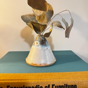 May include: A small, handmade ceramic vase with a speckled white and blue glaze, holding dried eucalyptus branches. The vase has a conical shape with a wide base. The vase is sitting on a teal book with the title "Encyclopedia of Furniture".