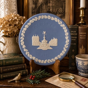 May include: A round, light blue ceramic plate featuring white relief decorations. The center depicts the Shaftesbury Memorial Fountain, flanked by buildings, with the text CHRISTMAS and 1971. A white holly leaf border encircles the central scene.