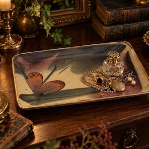 May include: A rectangular ceramic tray featuring a painted orange butterfly and dark green foliage on a cream-colored background. The tray holds a clear glass perfume bottle with a gold cap, a gold chain necklace, a gold ring, and pearl earrings.
