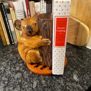May include: A ceramic koala bookend holding a book titled "Mastering the Art of French Cooking" by Julia Child and Bertholle Beck. The bookend is on a wooden base and is surrounded by a bookshelf with various cookbooks.