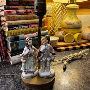 May include: A vintage table lamp with porcelain figurines of a man and woman in historical attire. The lamp has a brass base and a dark brown stem. The figurines are painted in pastel shades and are mounted on a decorative base.