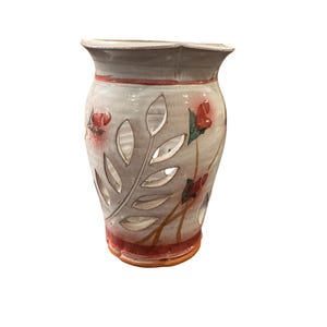 May include: A cream-coloured ceramic vase with cut-out leaf patterns and hand-painted red flowers. The vase has a scalloped rim and a red band around the top and bottom. The vase is a decorative item.