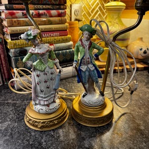 May include: Two antique porcelain lamps featuring figurines in period clothing. One lamp depicts a woman in a green and pink dress, the other a man in a green coat. Both lamps have gold-colored bases and are wired for electricity.