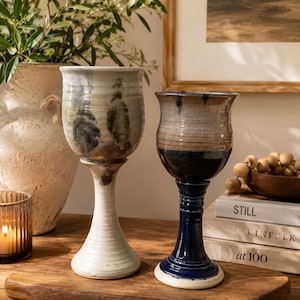 Puede incluir: Dos copas de cerámica con estética rústica. Una copa tiene una base blanca y una copa de color claro con detalles oscuros. La otra copa tiene una base azul oscuro y una copa marrón. Ambas están sobre una superficie de madera.