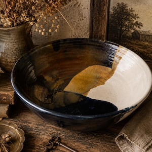 Puede incluir: Un cuenco de cerámica grande y redondo con un esmaltado de varios tonos que incluye secciones en negro profundo, ocre cálido y blanco crema. La superficie interior muestra un acabado liso y brillante con transiciones de color visibles, mientras que el borde exterior mantiene un tono oscuro constante, exhibiendo una estética artesanal con un diseño de cuenca abierta.