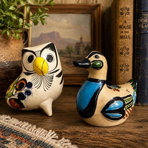 May include: Two ceramic figurines, an owl and a duck, featuring cream-colored bases with hand-painted details. The owl has a bright yellow beak and black eye markings. The duck displays a vibrant blue chest, a black head, and colorful wing patterns.