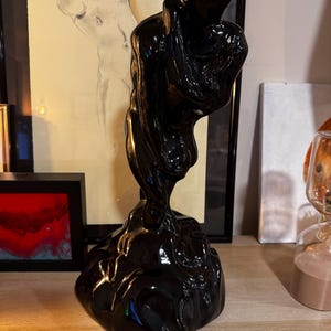 Könnte beinhalten: Eine glänzend schwarze Skulptur einer drapierten Figur, montiert auf einem strukturierten Sockel. Das Kunstwerk hat eine glatte, reflektierende Oberfläche und wird auf einem Holzregal ausgestellt. Die Skulptur ist ein modernes Kunstwerk.