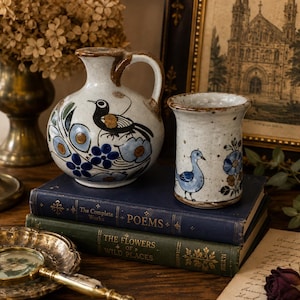 May include: A ceramic pitcher and a cylindrical cup, both featuring a speckled off-white glaze with hand-painted blue and black bird and floral motifs. The pitcher has a curved handle and a rounded body, while the cup has a flared rim.