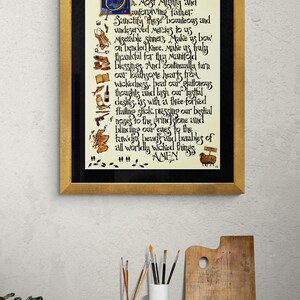 May include: A framed artwork with a gold-coloured frame and black matting. The artwork features calligraphy in black ink on a cream-coloured background. The text includes the word "Amen" at the bottom. The artwork is displayed on a wooden table with art supplies.